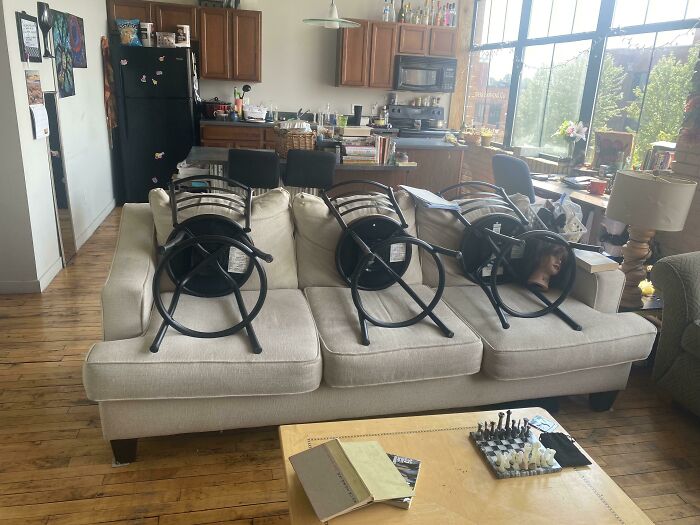 Living room with chairs upside down on couch, a chessboard on table, and bright windows in a messy roommate situation.