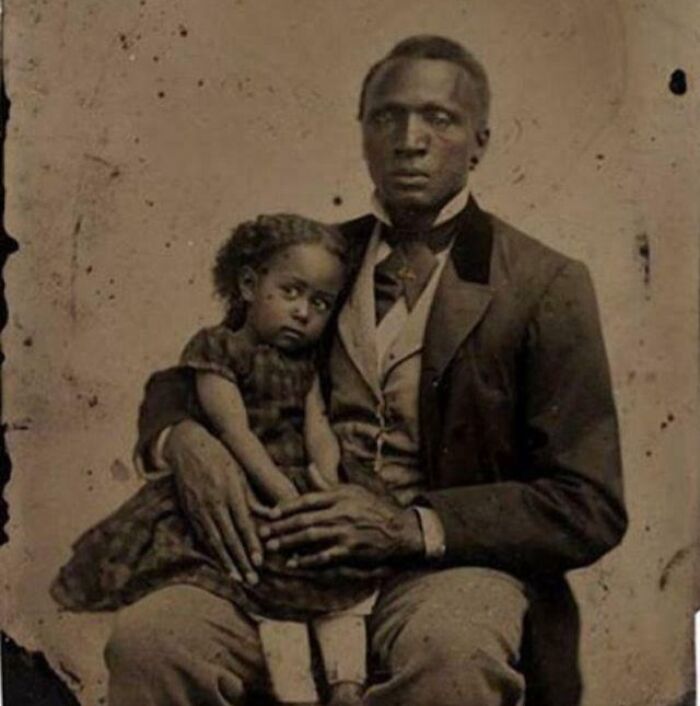 Black father holding young daughter, both dressed in 19th-century clothing in an interesting historical photograph.