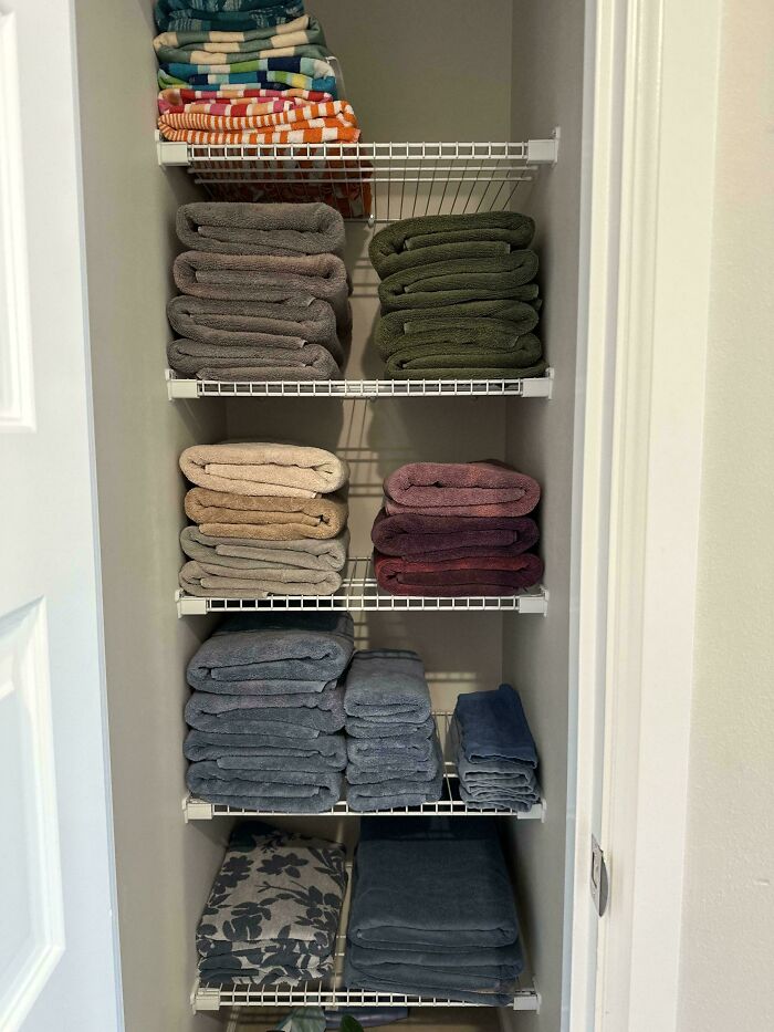 Neatly organized towels folded and stacked by color on white wire shelves in a small linen closet for order and tidiness.