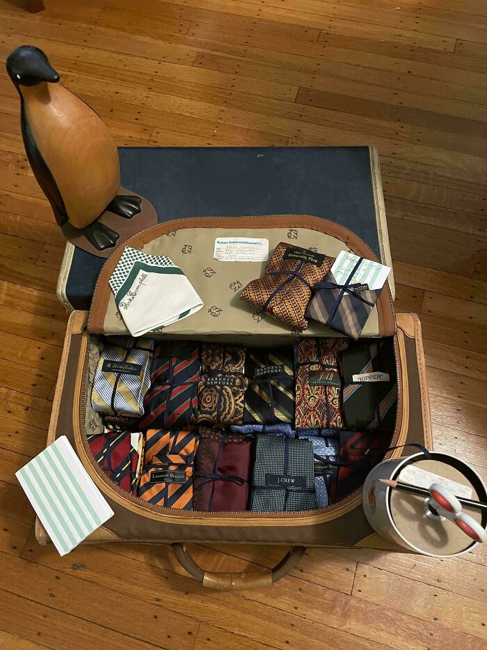 Neatly organized vintage suitcase with rolled patterned ties, handkerchiefs, and small wrapped packages on wooden floor.