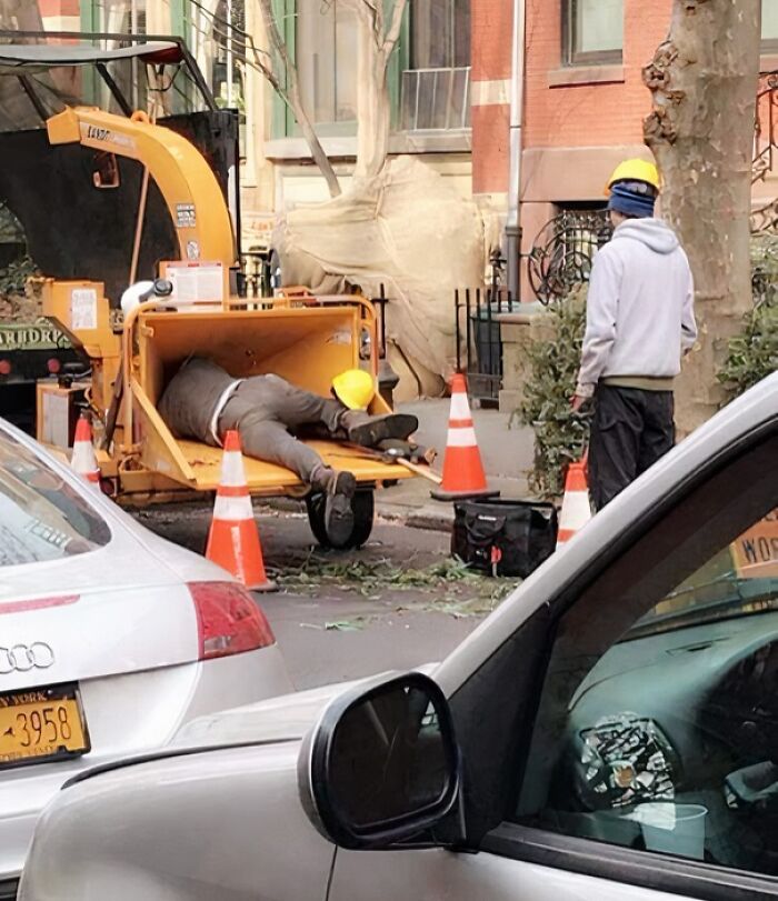 Worker dangerously leaning inside wood chipper machine with safety cones placed around on street, reckless work moment.