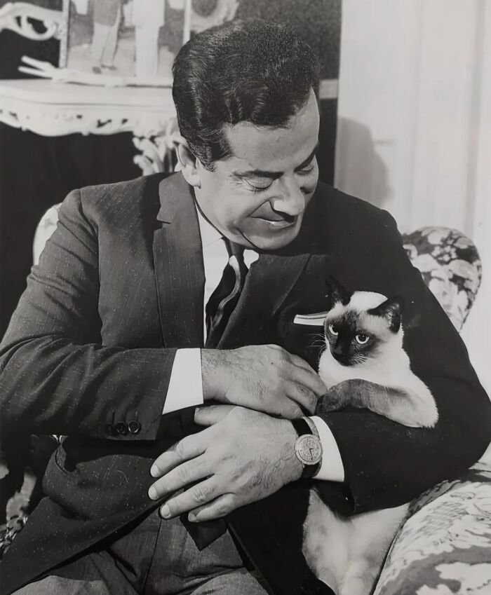 Man in a suit smiling while holding a Siamese cat, showcasing timeless feline charm in a vintage black-and-white photo.