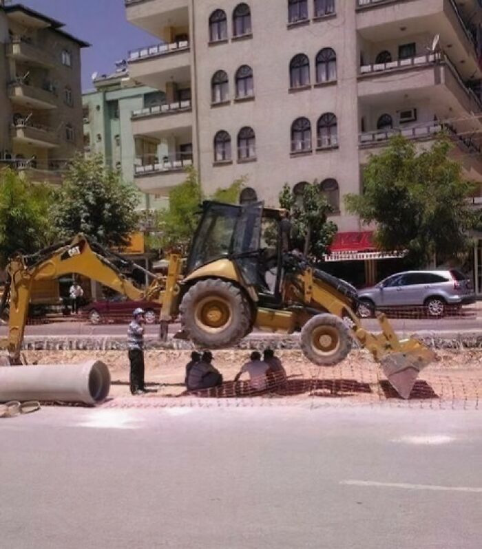 Construction workers sitting dangerously under a raised backhoe loader in a reckless work moment on a busy street.