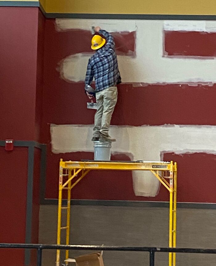 Worker standing recklessly on a bucket atop scaffolding without proper safety gear during a hazardous work moment.