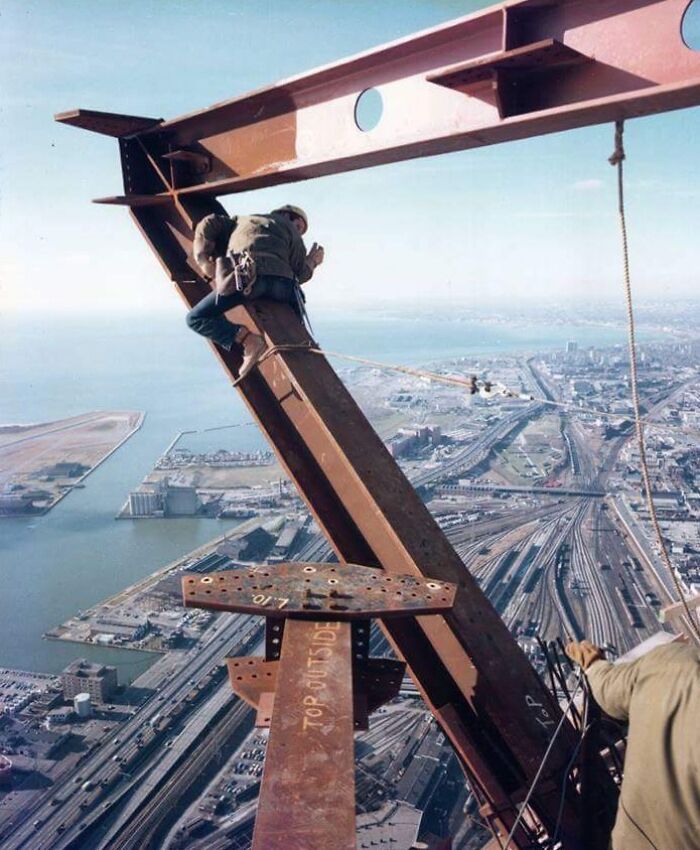 Worker climbing a steel beam high above city streets in a reckless work moment without visible safety gear.