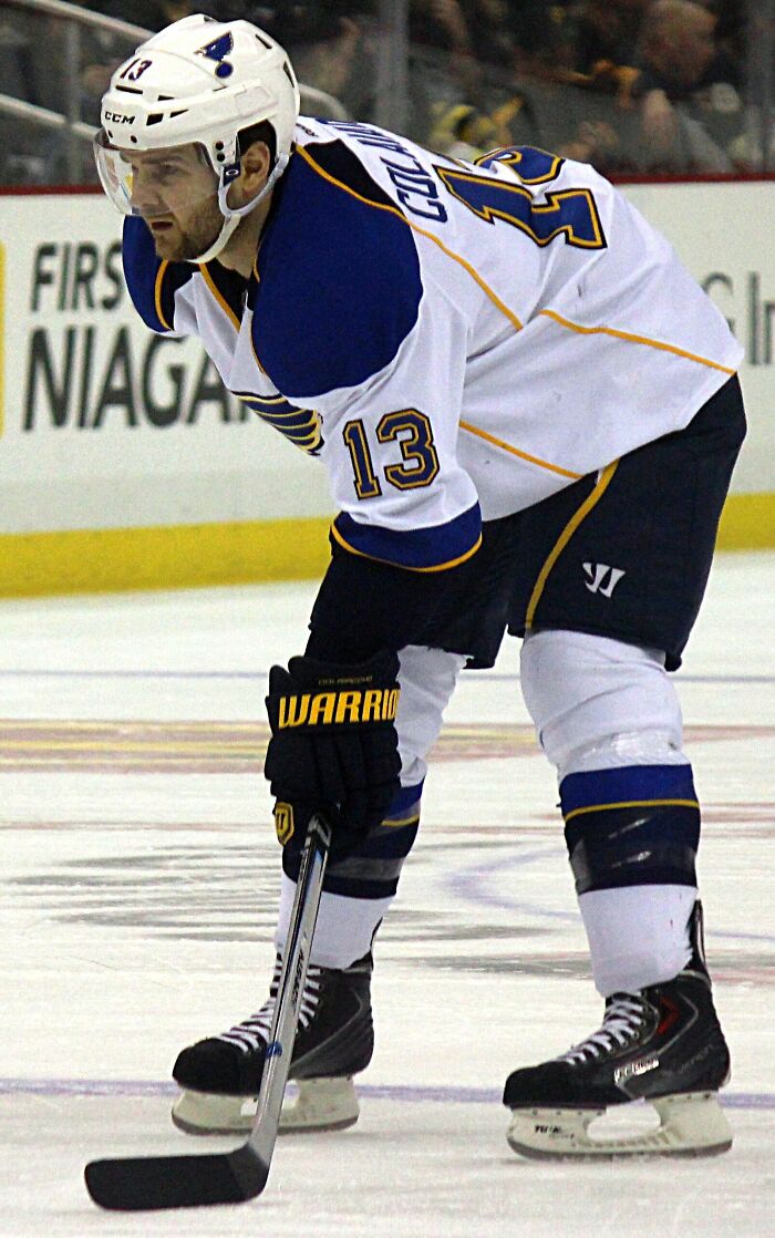 Carlo Colaiacovo in St. Louis Blues uniform, focused on the ice holding hockey stick during a game.