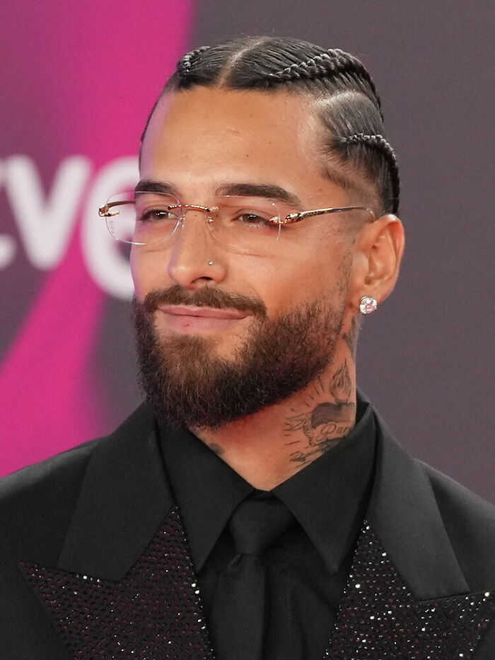 Maluma with braided hair and glasses, dressed in a black suit, at a formal event highlighting his career.