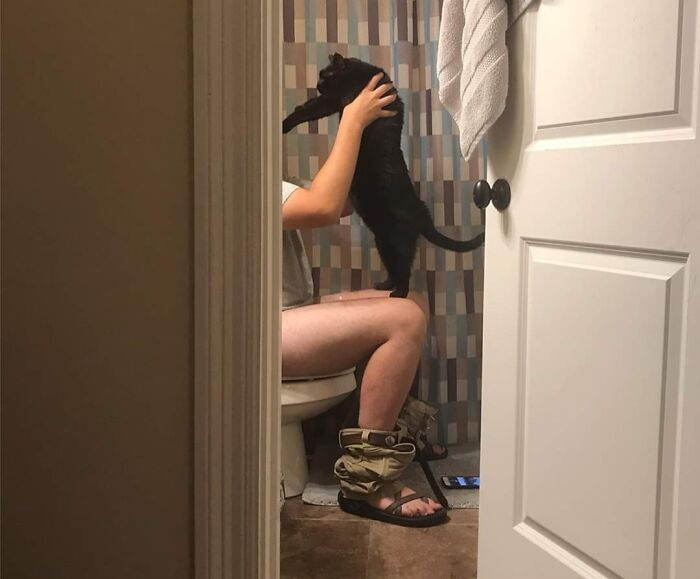Person sitting on toilet holding a black cat, showcasing a sweet human-animal bond in a bathroom setting.