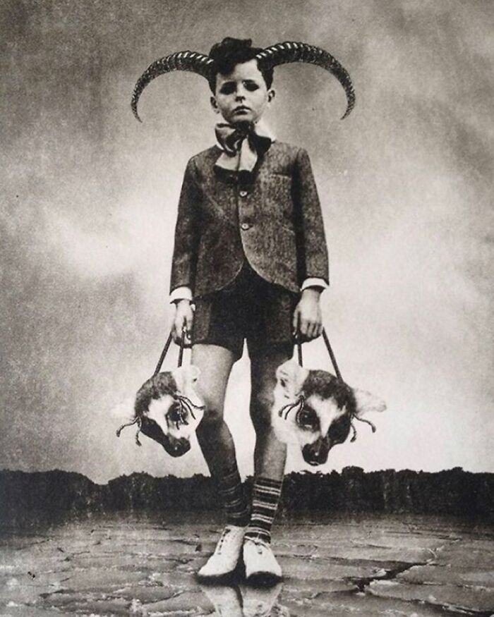 Boy with large curled horns holding two animal heads, vintage creepy black and white photo with odd eerie atmosphere.