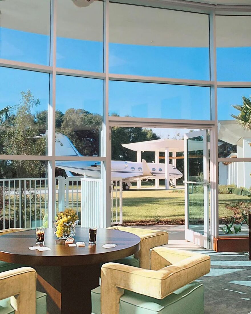 Luxurious modern celebrity house interior with large glass windows and private jet visible outside on the lawn.