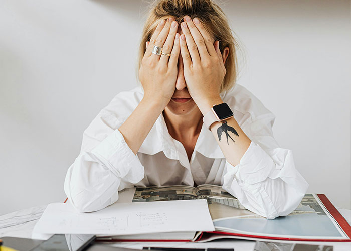 Young person with smartwatch and tattoo covering face, surrounded by papers and books, illustrating neurodivergent habits and challenges.
