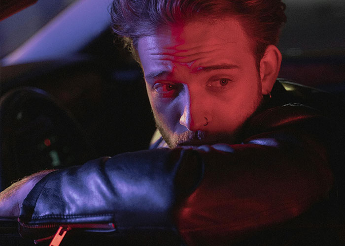 Man in leather jacket with nose ring, illuminated by red and blue light, reflecting on his wacky stories hoping to be believed.