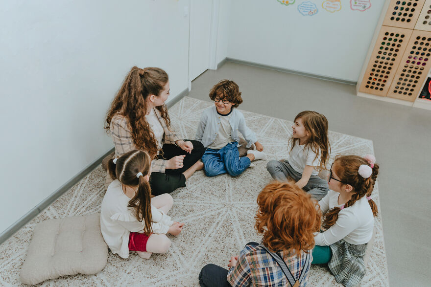 Group of kids and a teacher sitting in a circle on a carpet, engaging in paranoia questions and sharing secrets.