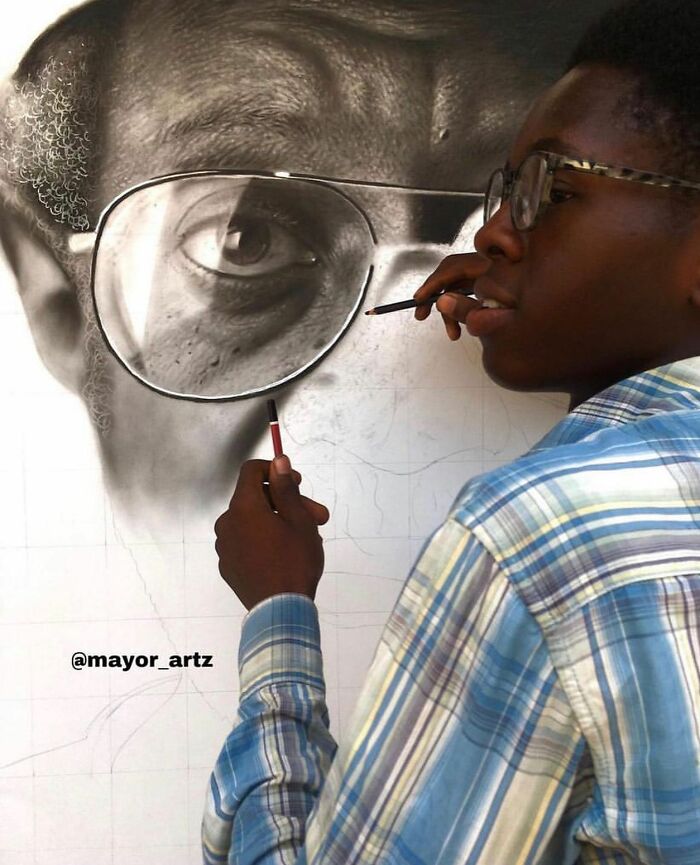 Artist creating hyperrealistic portrait using pencils, focusing on detailed eye and glasses in a behind-the-scenes photo.