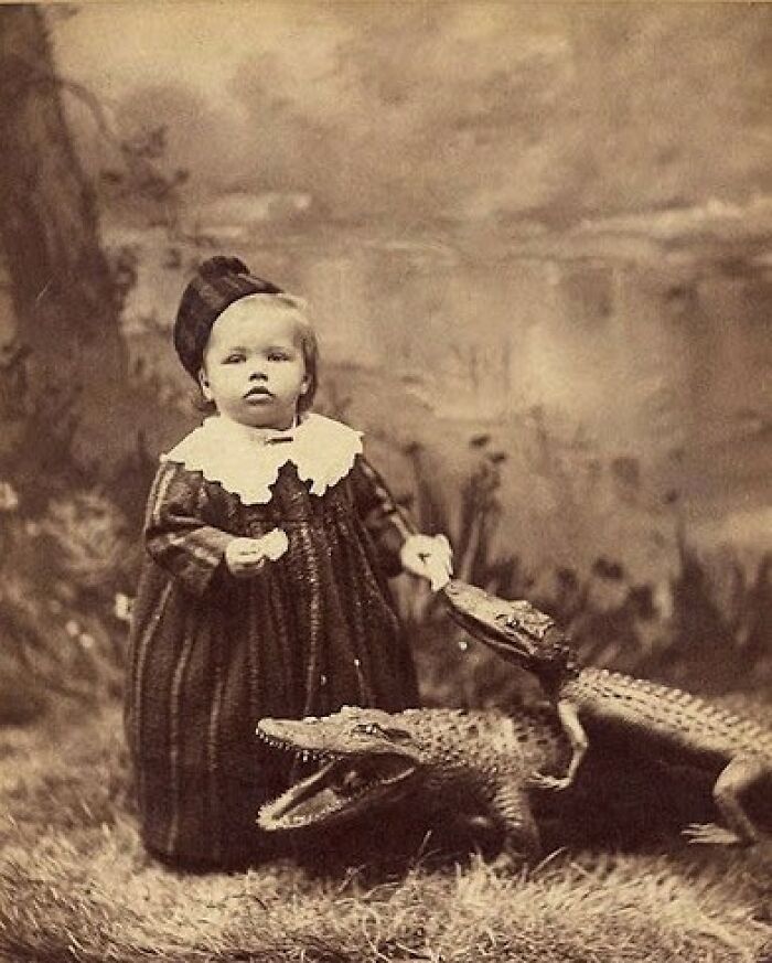 Vintage black and white photo of a child in old-fashioned clothes with two small crocodiles in a creepy odd scene.
