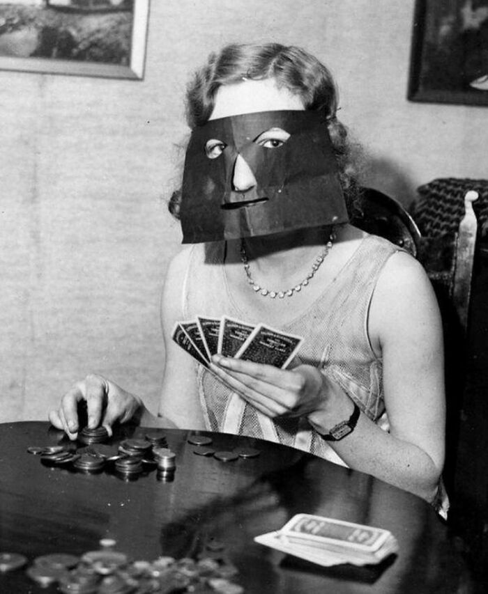 Woman wearing an odd black paper mask playing cards and stacking coins in an old black and white creepy photo.