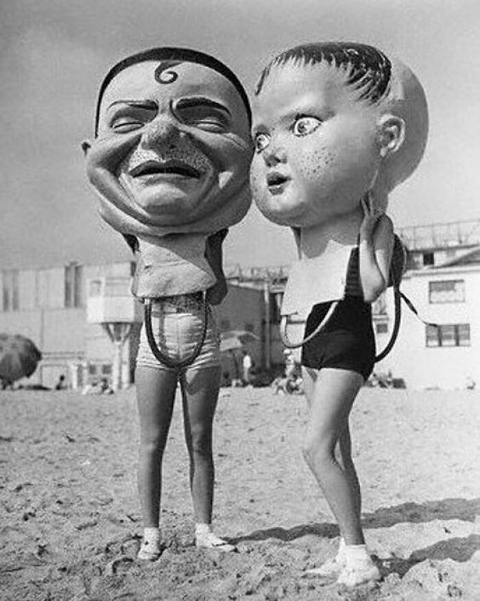 Two people standing on a beach wearing oversized odd and creepy black and white masks covering their heads.