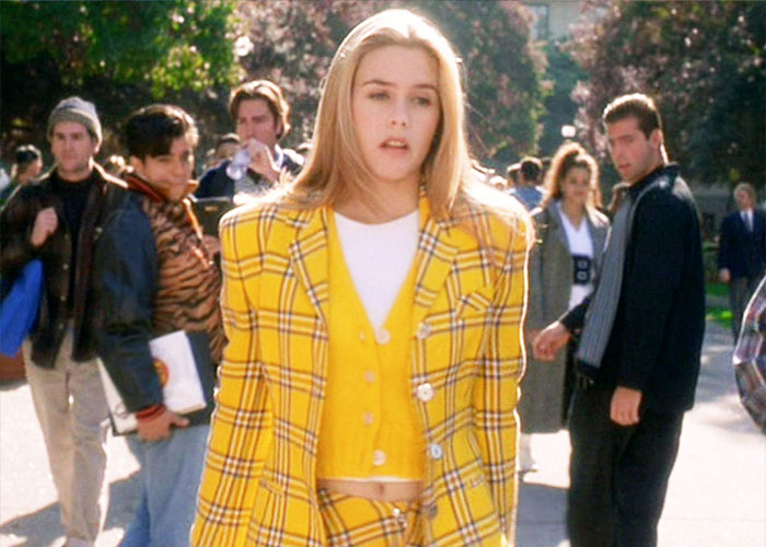 Alt text: Alicia Silverstone wearing a yellow plaid suit and vest walking outdoors among a group of people during the day. Alt text: Alicia Silverstone wearing a yellow plaid suit and vest walking outdoors among a group of people during the day.