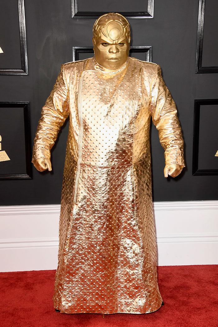 Person in an all-gold textured outfit and matching mask posing on the red carpet at the Grammys with wild fashion looks.