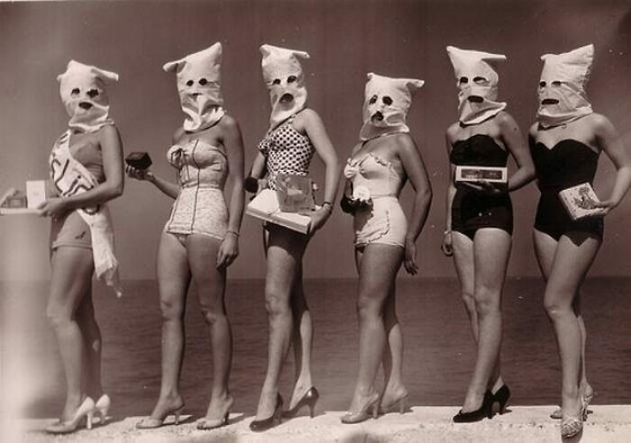 Group of women in vintage swimsuits and odd masks posing by the water in an old black and white creepy photo.