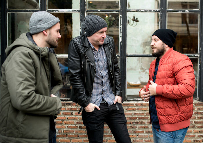 Three men dressed in winter jackets and beanies having a casual outdoor conversation about cultural differences in America.
