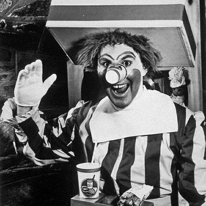 Old black and white creepy clown with a cup on his nose wearing striped outfit and waving his hand oddly.