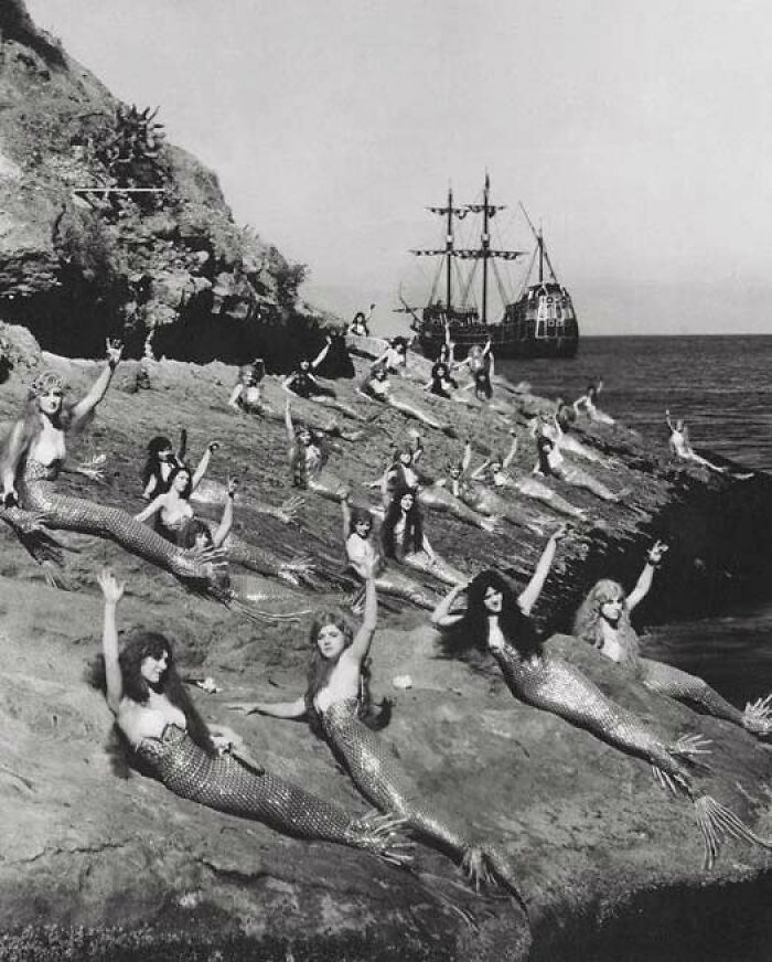 Black and white photo of women dressed as mermaids on rocky shore with old ship in background, odd and creepy vintage scene.