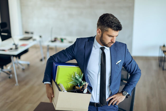 Man in a suit carrying a box of personal items, leaving an office in a satisfying well not my problem situation.
