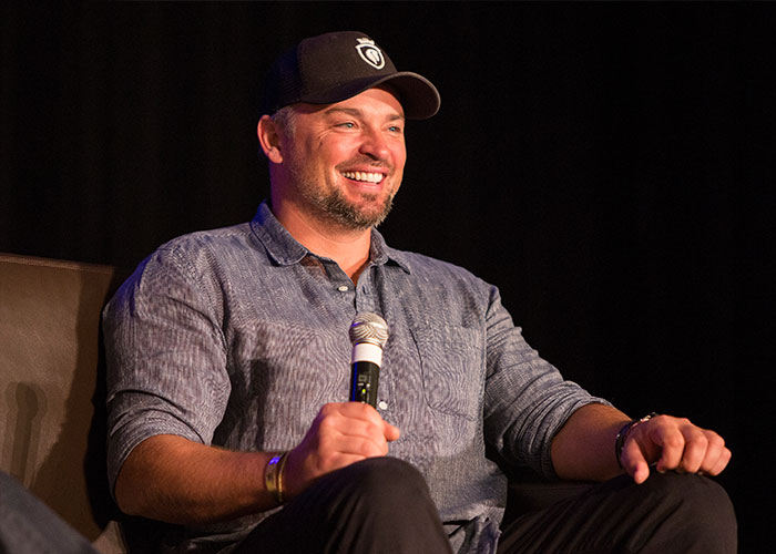 Man wearing a cap and casual shirt smiling while holding a microphone during a panel discussion about 2000s teen heartthrobs.