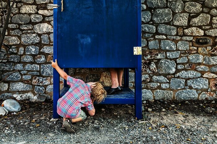 Child peeking under a blue booth in candid street photos capturing humanity, absurdity, and tender moments globally.