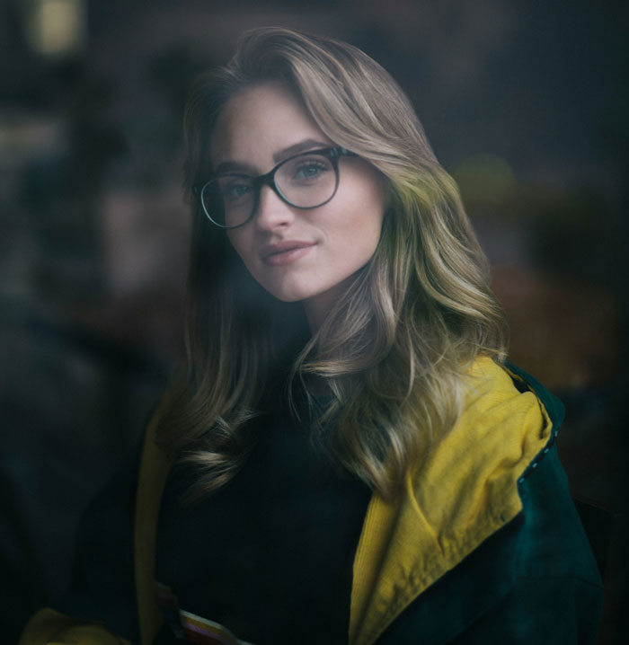 Young woman wearing glasses and a yellow jacket, looking thoughtful and smart in a dimly lit room.