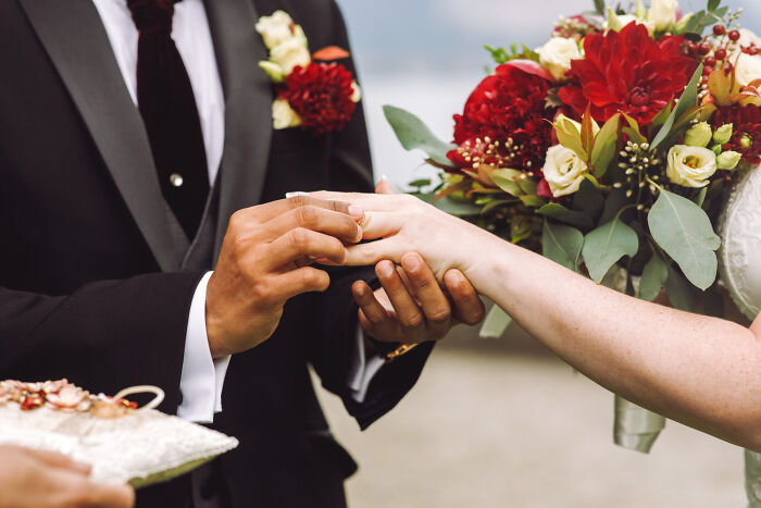 Groom placing wedding ring on bride’s hand during ceremony with bridal bouquet featuring red and white flowers.