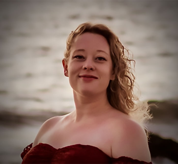 Young woman with curly hair wearing a red off-shoulder dress by the water, related to family of mom slain by ICE agent. Young woman with curly hair wearing a red off-shoulder dress by the water, related to family of mom slain by ICE agent.