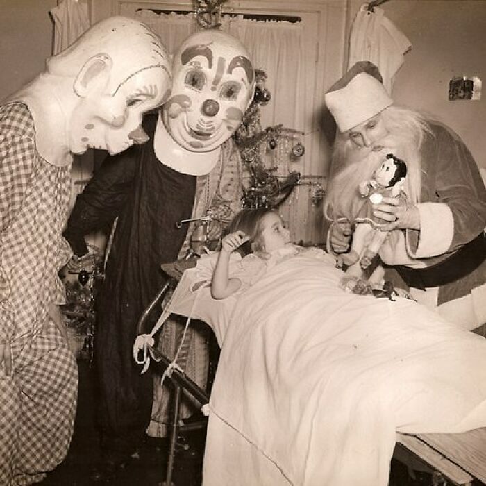 Child in hospital bed visited by adults in creepy clown and Santa costumes in old black and white photo.