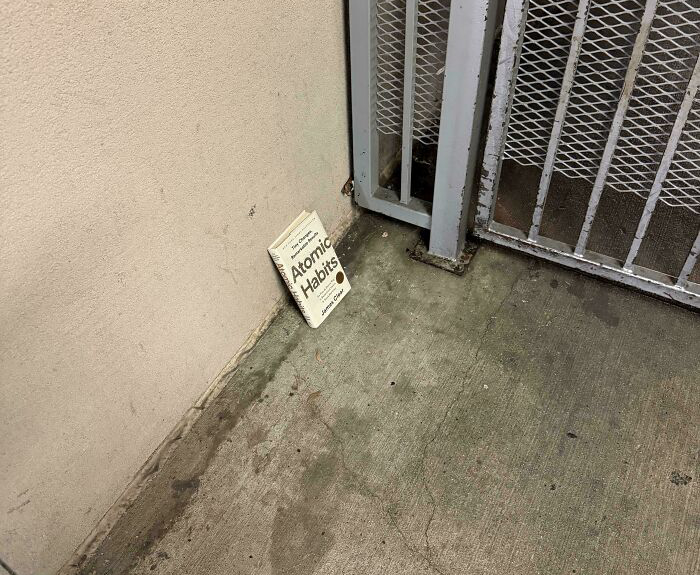 Paperback copy of Atomic Habits left abandoned on dirty concrete floor next to metal gate in confusing pictures funny for no clear reason