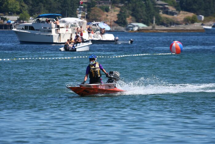 Person riding a small motorized boat on the water during a strange tradition event with boats and spectators nearby.