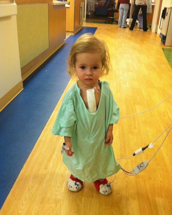 Toddler in hospital gown with medical tubes attached, standing in a hospital corridor, showing a fascinating and unnerving medical scene.
