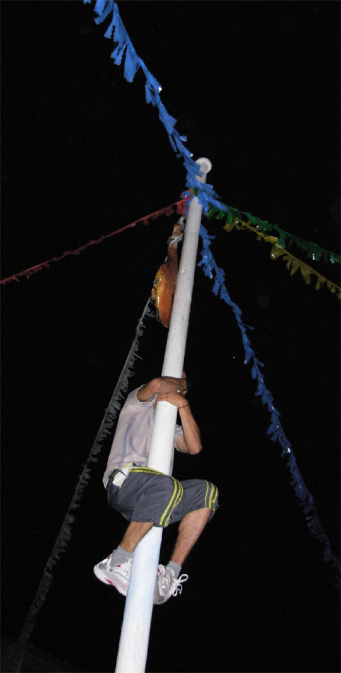 Man climbing a tall pole decorated with colorful flags during a strange tradition from around the world at night.