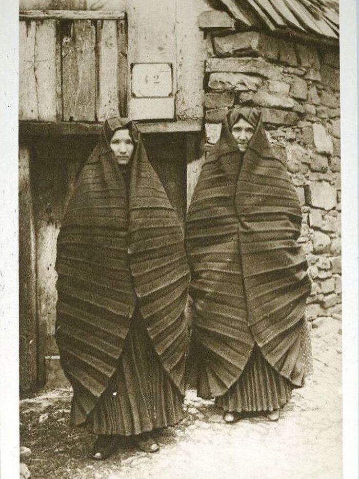 Two women wrapped in traditional striped shawls standing outside a rustic stone building in history photographs.