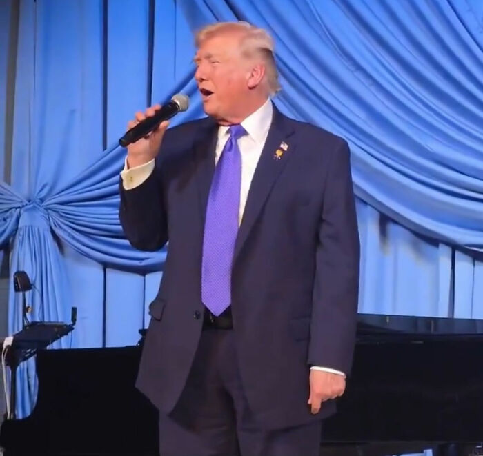 Man in a suit holding a microphone on stage at a Mar-a-Lago event with blue draped curtains in the background. Man in a suit holding a microphone on stage at a Mar-a-Lago event with blue draped curtains in the background.