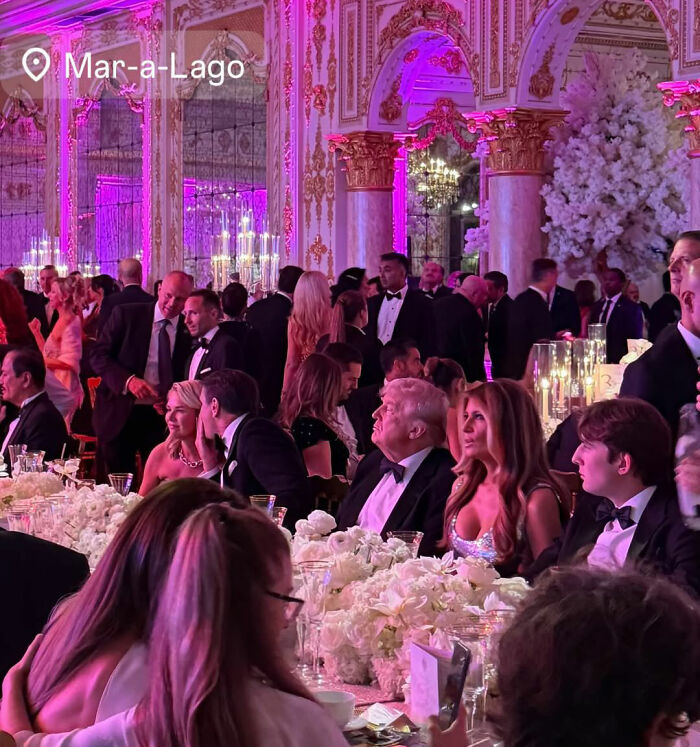 Guests dressed in formal attire at a lavish event inside Mar-a-Lago with elegant decor and pink lighting. Guests dressed in formal attire at a lavish event inside Mar-a-Lago with elegant decor and pink lighting.