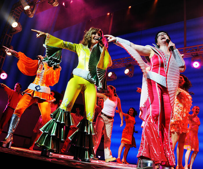 Male Karen shouting at Mamma Mia Broadway performance as colorful 70s-costumed cast dances onstage