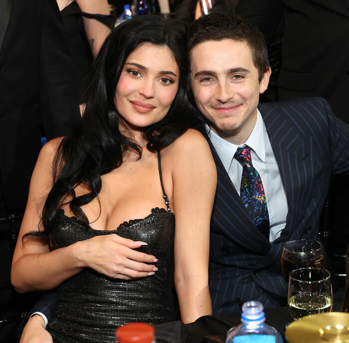 Timothée Chalamet smiling in a pinstripe suit next to a woman in a black dress at a formal event gathering. Timothée Chalamet smiling in a pinstripe suit next to a woman in a black dress at a formal event gathering.