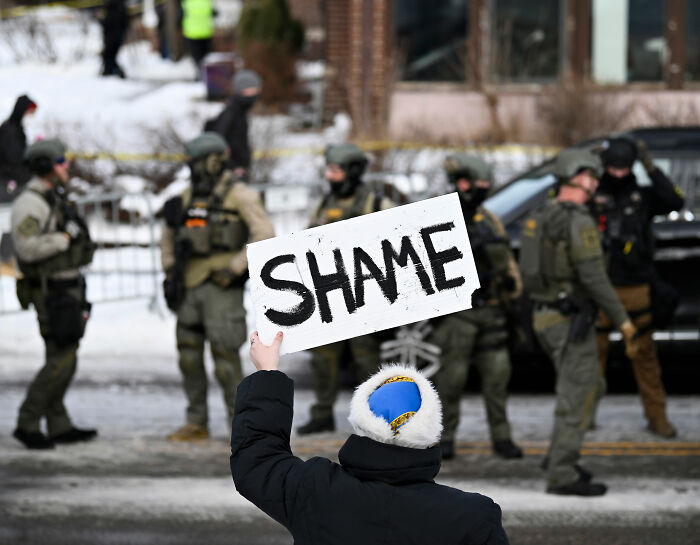 Protester holding a shame sign facing ICE agents in tactical gear during a public demonstration on a snowy street. Protester holding a shame sign facing ICE agents in tactical gear during a public demonstration on a snowy street.