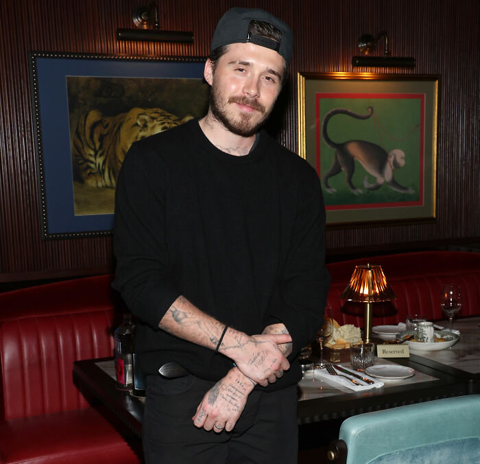 Brooklyn Beckham wearing a black cap and sweater, standing in a restaurant during a casual evening.