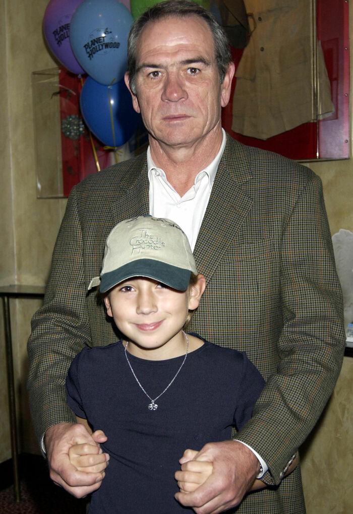 Tommy Lee Jones standing behind and holding hands of a young girl wearing a cap at an indoor event.