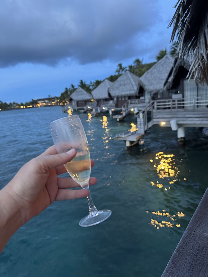 Hand holding a champagne glass over turquoise blue water with overwater bungalows in French Polynesia at dusk.