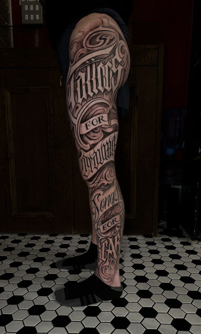 Full leg tattoo with detailed black and gray lettering and intricate swirling designs on skin.