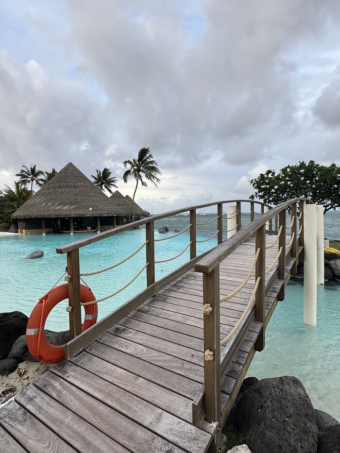 Wooden bridge over turquoise water near thatched huts and palm trees in French Polynesia with shades of blue sky.