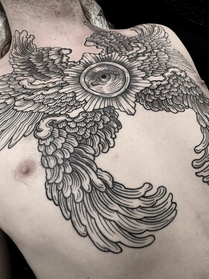 Black and grey tattoo of a detailed eye with wings covering a person’s upper chest, showcasing cool tattoo design.